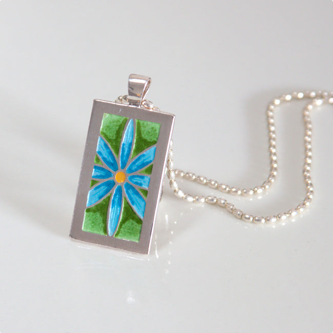 Enamel pendant necklace with blue daisy, yellow center, green background, on fine silver. Hung on silver chain.